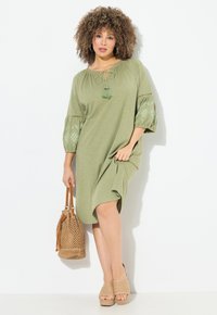 Oliwkowa sukienka do kolan z teksturowanymi rękawami, dekoltem ze sznurkiem i frędzlami. Połączona z plecioną torbą i beżowymi platformami.
