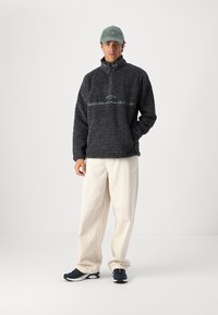 Fleece pullover in dark gray with a half-zip collar and textured finish. Paired with loose-fitting cream pants and black sneakers.