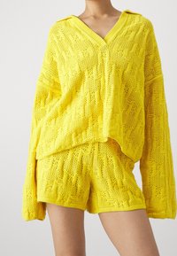 Stickad gul tröja och shorts-set med en texturerad, öppen struktur, med V-ringning och vida ärmar. Båda delarna har en avslappnad passform.