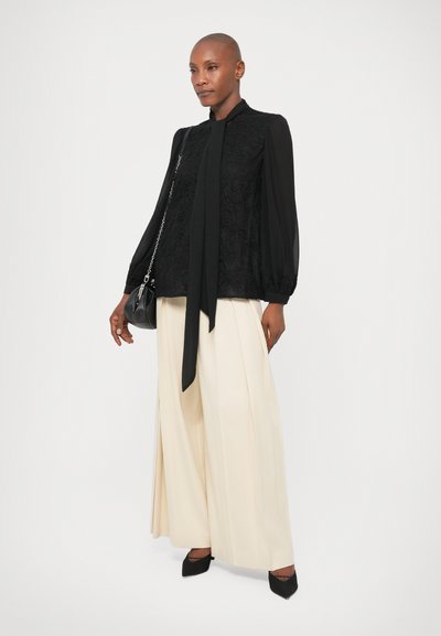 Melnas mežģīņu blūze ar kakla saitēm un pūkainām piedurknēm, apvienota ar krēmkrāsas platām biksēm un melniem kurpēm ar izteikto purngalu. Mazs melns rokassomas.