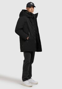 Hombre vestido con una chaqueta de invierno con capucha negra, gorra de béisbol negra, jeans oscuros y zapatillas blancas de pie contra un fondo sencillo.