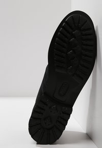 Suola di scarpa nera con una superficie in gomma testurizzata e scanalature profonde per la trazione. Presenta un lato liscio con dettagli di cucitura.