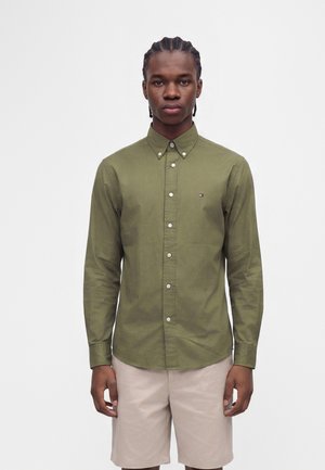 Junger Mann mit geflochtenem Haar, der ein olivgrünes Hemd mit Knöpfen und beige Shorts trägt, steht vor einem schlichten hellen Hintergrund.