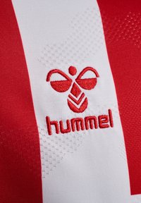 Rød og hvid sports-trøje med et struktureret design, prydet med et rødt broderet logo af "hummel" og et stiliseret bi-motiv.