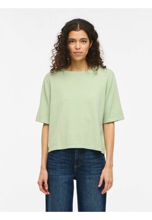 Kvinde med krøllet mørkt hår iført en lysegrøn kortærmet top og blå jeans, stående mod en ensartet hvid baggrund.