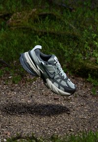 Scarpa sportiva grigia e verde con tomaia in rete, sovrapposizioni testurizzate, suola spessa e accenti neri. Fluttua sopra una superficie ghiaiosa.