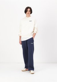 Cream sweatshirt with small black logo, paired with navy soft pants. Shoes are white with red stripes. Simple, relaxed fit design.