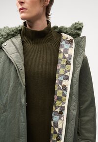 Giacca trapuntata verde oliva con cappuccio testurizzato, caratterizzata da un rivestimento patchwork multicolore e bottoni marroni, indossata sopra un maglione a coste scuro.