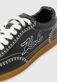 Black leather sneaker featuring white stitching and laces, with a tan rubber sole. Embroidered "Karl" logo on the side.