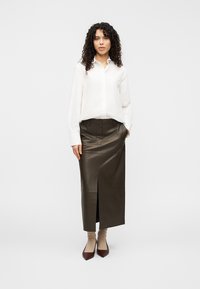 Καφέ δερμάτινη φούστα με υψηλή σκίσιμο, σε συνδυασμό με λευκό πουκάμισο με κουμπιά. Το σύνολο διαθέτει λεία υφή και εφαρμοστή γραμμή.