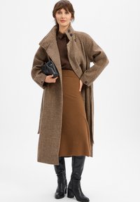 Frau in braunem kariertem Mantel, brauem Rock, dunkelbraunem Hemd, schwarzen High-Heels, die eine kleine schwarze Clutch vor einem weißen Hintergrund hält.