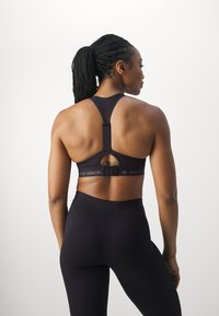 Schwarzer Sport-BH mit Racerback-Design und verstellbarem Hakenverschluss. Besitzt ein Cutout und ein Logo-Band. Wird mit passenden schwarzen Leggings getragen.