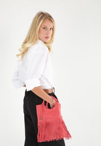 Koralowa skórzana torebka z frędzlami, z górnym uchwytem i okrągłym wycięciem. Modelka ma na sobie białą koszulę i czarne spodnie.