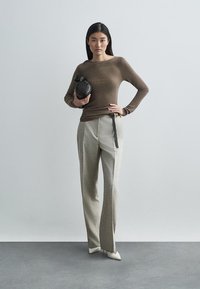 Smeđi uski džemper s dugim rukavima u kombinaciji sa svijetlosivim hlačama s visokim strukom. Model drži okruglu crnu torbu; izgled upotpunjuju šiljaste bijele štikle.