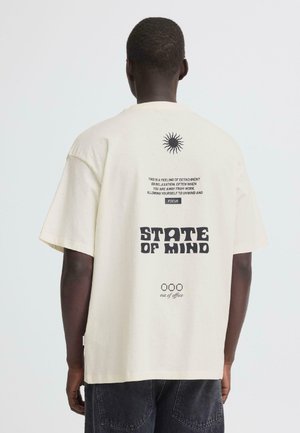 Mężczyzna w beżowej koszulce z czarną grafiką słońca i napisami "STATE OF MIND" oraz "out of office" na plecach, w połączeniu z ciemnymi dżinsami.