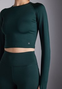 Langermet kort topp og høyt liv leggings i mørkegrønn stoff. Glatt tekstur med minimale sømmer og en liten logo på toppen.