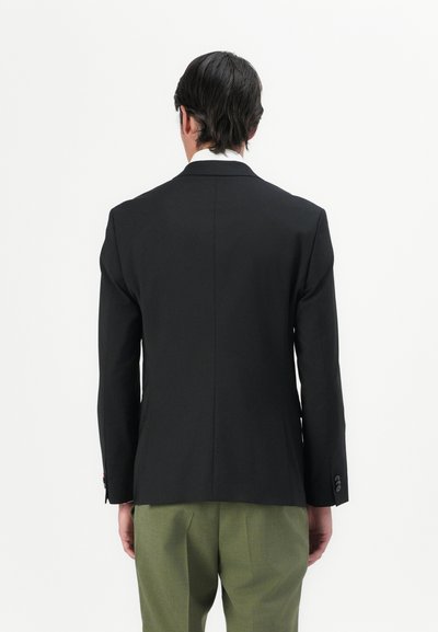 Blazer noir cintré avec une texture lisse, fente unique à l'arrière et poignets boutonnés, associé à un pantalon vert clair.