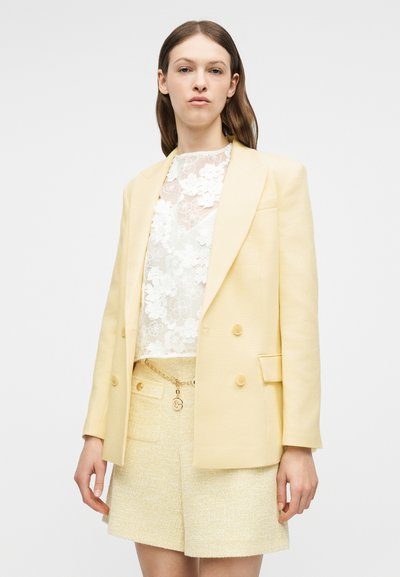 Femme portant un blazer jaune clair sur un haut en dentelle florale blanche et un short texturé avec un détail chaîne sur fond blanc.