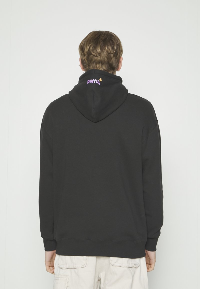 Personne aux cheveux courts portant un sweat à capuche noir avec le texte violet « puma » au dos de la capuche, assorti à un pantalon cargo clair.