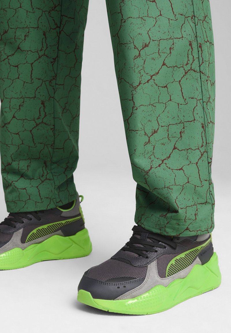 Svarta och grå sneakers med ljusgröna accenter och en texturerad sula, kombinerat med gröna byxor som har ett sprucket brunt mönster.