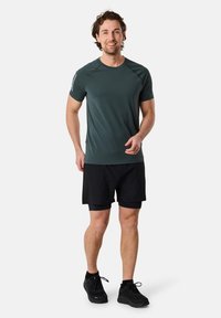 Donkergroene sportshirt met korte mouwen en een gladde textuur, gecombineerd met zwarte shorts en zwarte sportschoenen. Heeft een eenvoudig ontwerp.