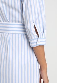 Abito a righe blu e bianche, caratterizzato da maniche arrotolate con chiusura a bottone e spacchi laterali, realizzato in tessuto leggero con una texture liscia.