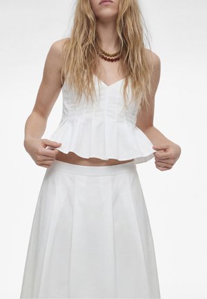 Femme aux longs cheveux blonds ondulés porte un haut blanc sans manches à basque et un pantalon large assorti, tenant l’ourlet du haut avec les deux mains.