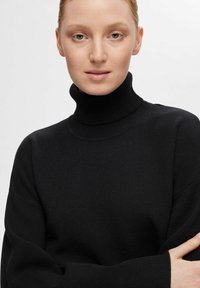 Črna turtleneck pulover iz mehkega, teksturiranega pletenja z visokim ovratnikom in spuščenimi rameni; ima sproščen kroj in rebraste manšete.