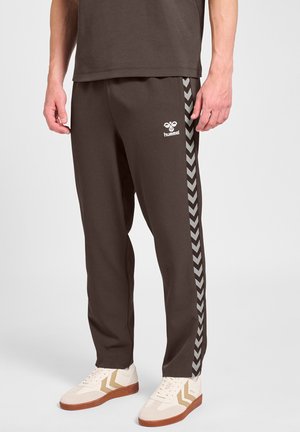 Mężczyzna noszący ciemnobrązowe spodnie sportowe Hummel z paskami w kształcie jodełki po bokach oraz białe sneakersy z brązowymi akcentami.