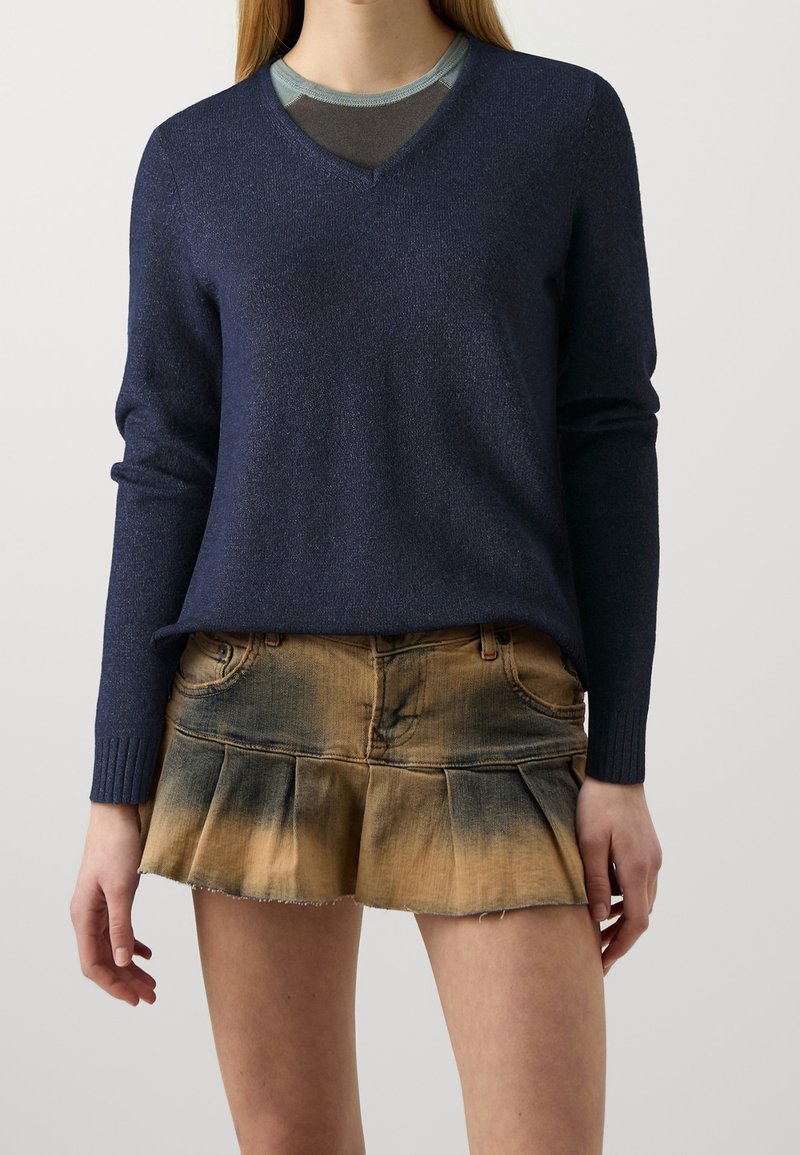 Femme portant un pull bleu marine à col en V par-dessus un top gris, associé à une jupe courte plissée en denim avec un motif marron et noir délavé.