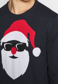 Marinblå jultröja med en stickad tomteansiktet som har en röd hatt, svarta solglasögon, röd näsa och vita skäggdetaljer.