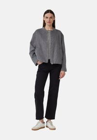 Veste bomber grise en laine avec fermeture éclair, dotée de deux poches avant et d'une chemise rayée en dessous, associée à un pantalon noir et des baskets blanches.