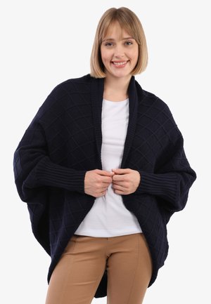 Marineblauer, gestrickter Wickel-Cardigan mit einem Diamantmuster, gerippten Ärmeln und einem offenen Design. Kombiniert mit einem weißen T-Shirt und beigen Hosen.