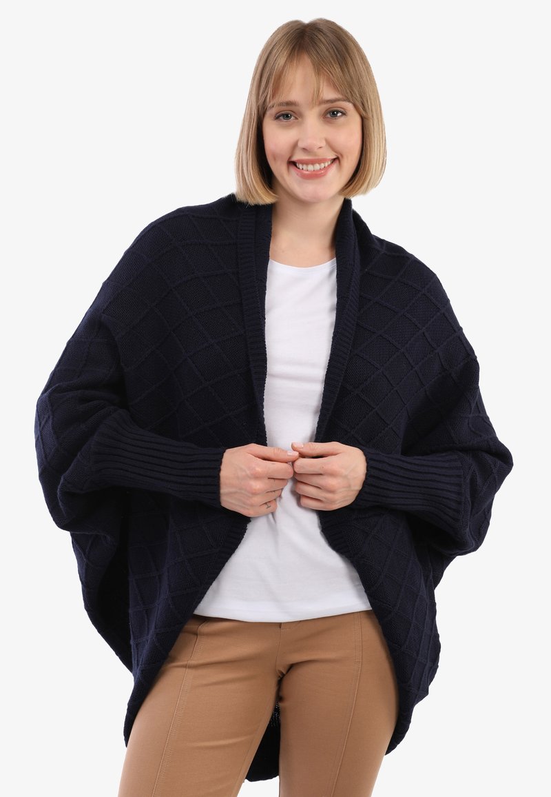 Marineblauer, gestrickter Wickel-Cardigan mit einem Diamantmuster, gerippten Ärmeln und einem offenen Design. Kombiniert mit einem weißen T-Shirt und beigen Hosen.