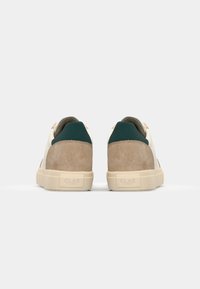 Sneaker con pannelli in suede e pelle; corpo crema, tacco marrone chiaro, dettagli verdi e suola beige. Logo del marchio sul tacco. Design minimalista.