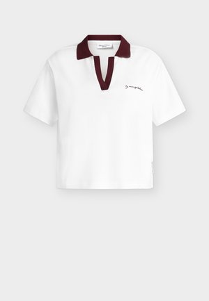 Άσπρο polo μπλουζάκι με κοντό μανίκι, σκούρο κόκκινο γιακά και πατιλέτα, μικρό μαύρο λογότυπο με γράμματα στο αριστερό στήθος και ετικέτα στη ραφή του πλευρού.