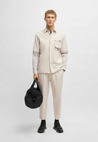 Beige shirt met korte mouwen en borstzak, gecombineerd met lichtbeige broek. Houdt een zwarte ronde tas vast en draagt zwarte sneakers.