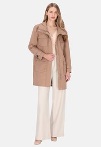 Beige nylonparka med hætte, flere forlommer, lynlås og trykknapper. Båres over en lysefarvet top og lyse bukser.