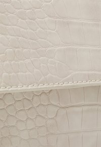 Kremowa skóra z tłoczeniem imitującym skórę krokodyla, o teksturowanej powierzchni, z delikatnym połyskiem i drobnym przeszyciem wzdłuż krawędzi.