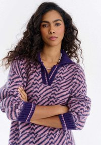 Kobieta z kręconymi włosami, ubrana w sweter w fioletowo-różowy wzór jodełki, z założonymi na siebie ramionami i koralikowym naszyjnikiem na białym tle.