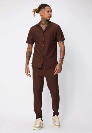 Chemise marron à manches courtes et pantalon tapered assorti, avec des rayures verticales. Porté avec des baskets de couleur claire, mettant en avant des détails de design tendance.