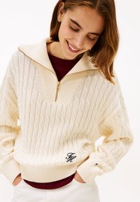 Kremfarget strikkegenser med halvlengd glidelås, med et teksturert diamantmønster og ribbestrikkede mansjetter. Pyntet med en brodert logo.