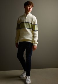 Pull-over en maille blanche avec une demi-fermeture éclair et des rayures horizontales vert olive et bleu marine, associé à un pantalon bleu foncé et des baskets blanches.