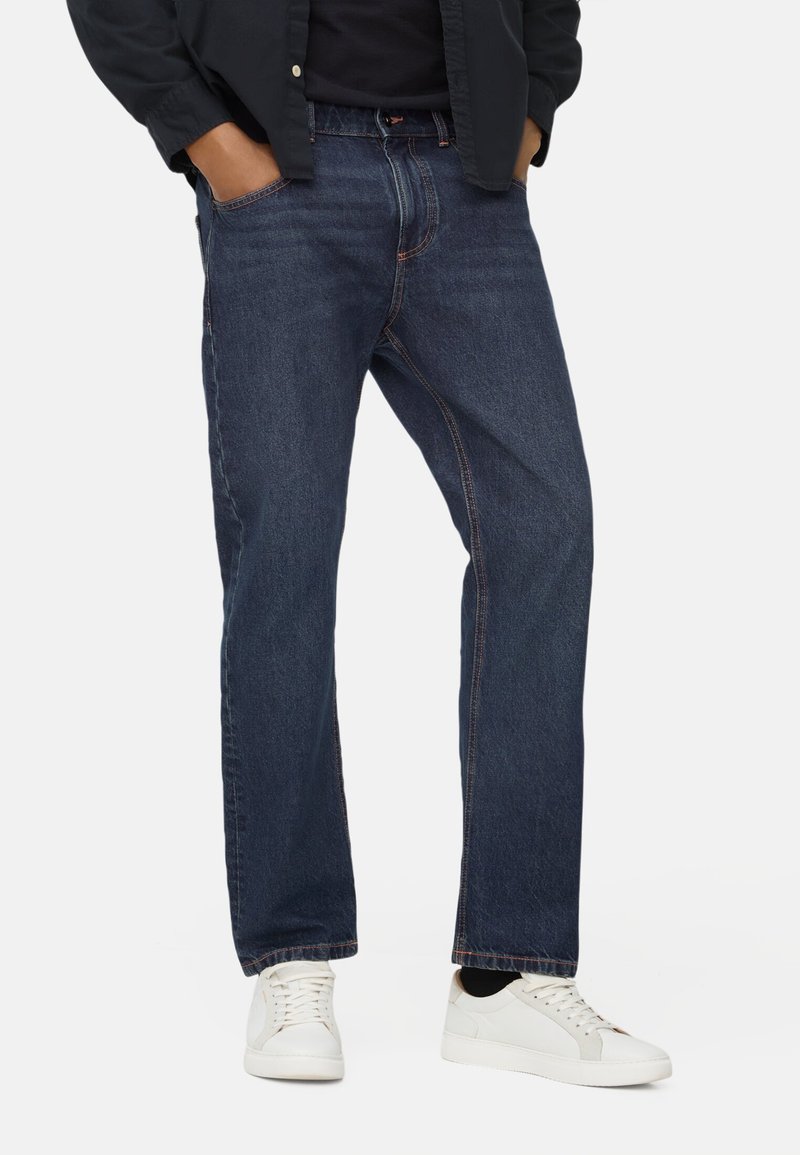 Donkerblauwe denim jeans met een recht model, voorzien van oranje stiksels en twee voorkeurszakken. Gepaard met witte sneakers.