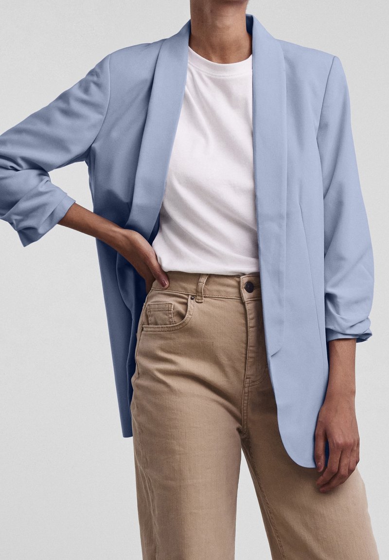 Personne portant un blazer bleu clair sur un t-shirt blanc et un pantalon beige taille haute, debout avec une main sur la hanche devant un fond uni.