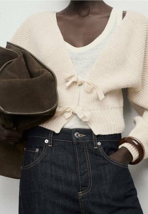 Personne portant un cardigan court en tricot crème noué avec des rubans, un haut blanc, un jean taille haute foncé, un bracelet marron, tenant un grand sac marron.