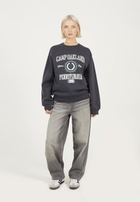 Donna con capelli biondi corti che indossa un felpa scura con la scritta "Camp Oakland Pennsylvania", jeans grigi larghi e sneakers bianche, in piedi su uno sfondo bianco.
