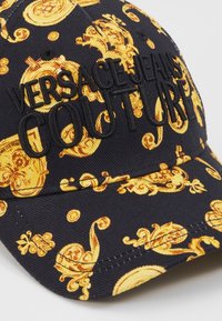 Casquette noire avec des motifs baroques dorés ornés et un texte noir en relief "Versace Jeans Couture" sur le panneau avant et la visière.