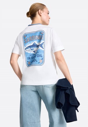 Woman with hair in bun wearing white t-shirt with blue sardines in olive oil label graphic on back, holding navy jacket, light jeans.