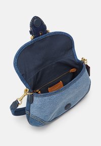 Petit sac bandoulière en jean bleu avec rabat ouvert, montrant la doublure intérieure bleu marine, une poche zippée, un patch logo en cuir et des accessoires dorés.